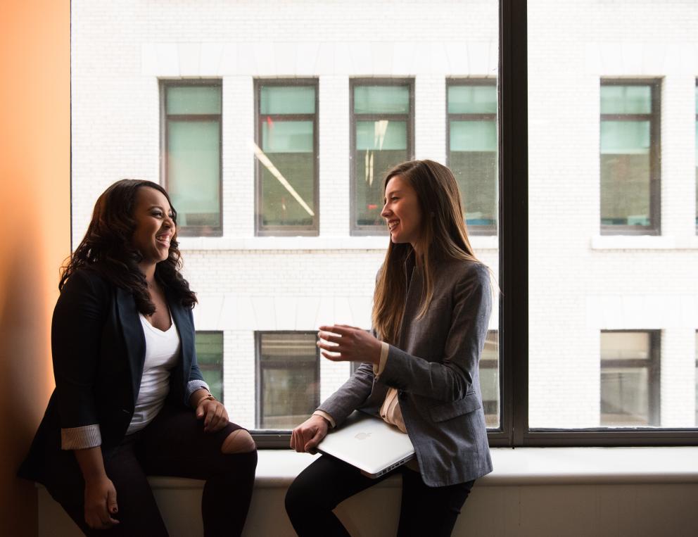 Two women are shown in business clothes, sitting on a windowsill and chatting.